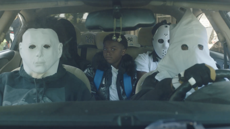A group of people ride in a car, four of them wear scary masks while one little boy is unmasked.